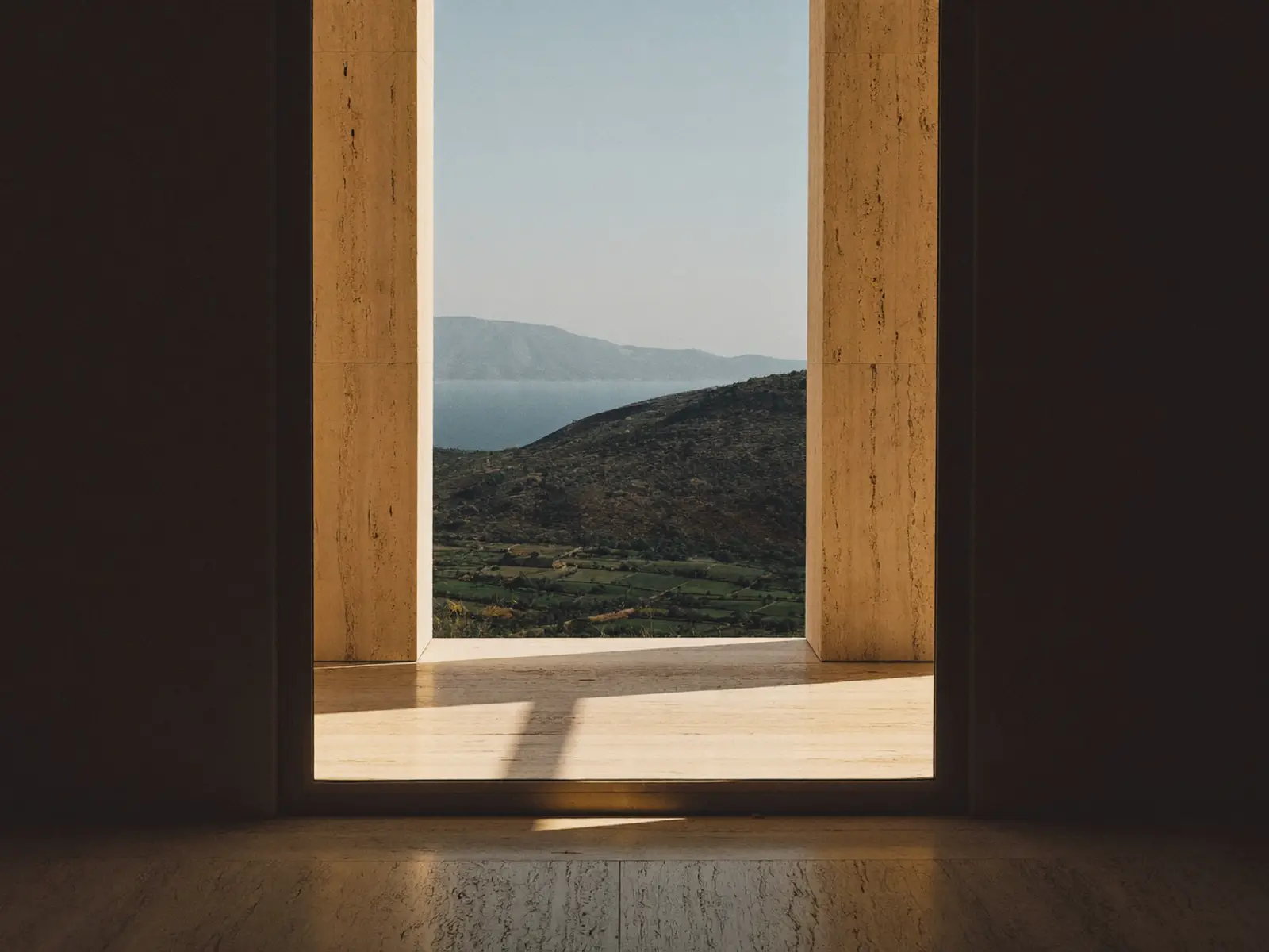 A travertine portal in a dark room, opening onto a green hillside and the sea